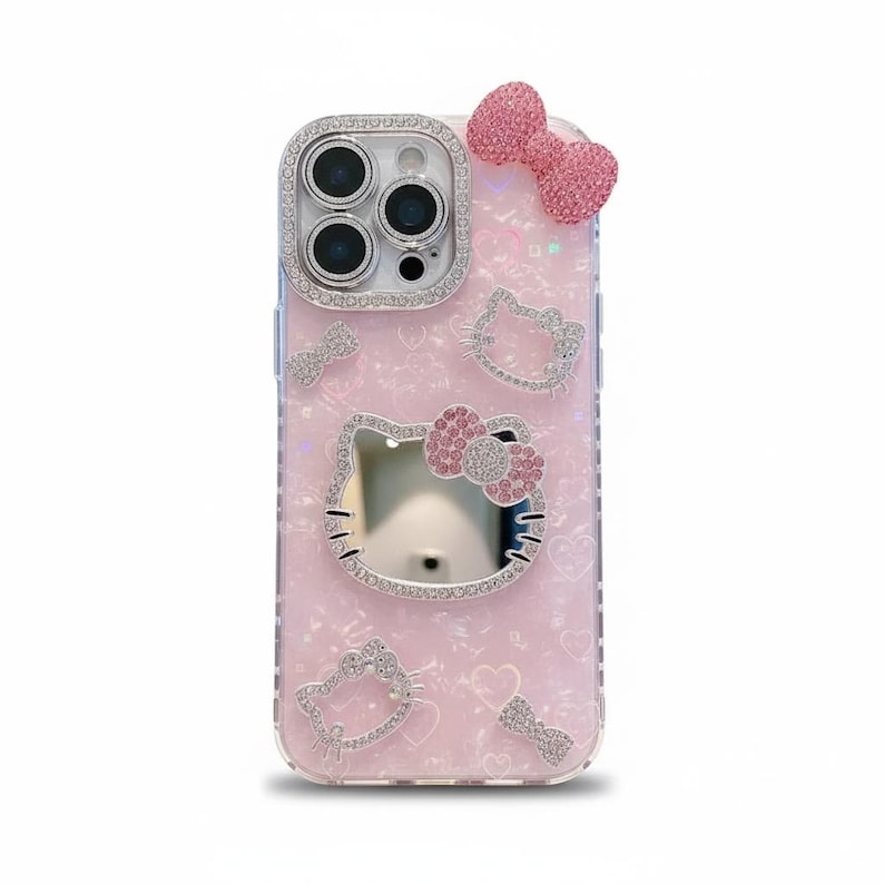 May include: Pink phone case with a Hello Kitty design. The case features a bow, Hello Kitty face, and other decorative elements, all encrusted with rhinestones. The camera lens is surrounded by a silver rhinestone border.