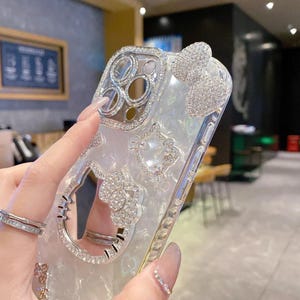 May include: A clear phone case with a silver frame and embellished with rhinestones. The case features a bow, a Hello Kitty design, and a ring holder. The camera lens area is also decorated with rhinestones.