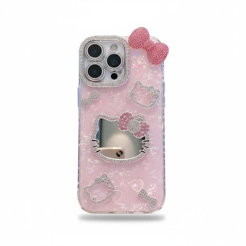 May include: Pink phone case with a Hello Kitty design. The case features a bow, heart, and Hello Kitty face embellishments, all adorned with rhinestones. The camera lens is surrounded by a rhinestone border.