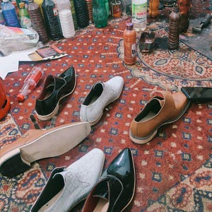 May include: Assortment of men's dress shoes in various colors, including black, brown, and white, arranged on a patterned red rug. The shoes have different textures and styles, with a focus on leather and a glossy finish. Bottles and containers are in the background.