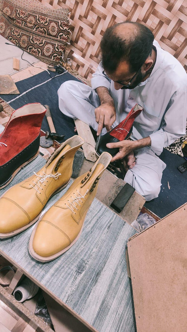 Peut inclure: Un cordonnier travaille sur une paire de bottes en cuir. Les bottes sont de couleur beige et rouge, avec des lacets blancs. Le cordonnier utilise un couteau pour couper le cuir. D'autres outils et mat&eacute;riaux sont visibles.