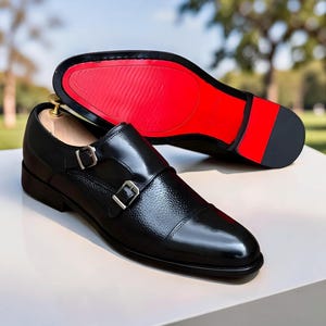 Peut inclure: Chaussures de ville noires en cuir avec double bride et boucles argent&eacute;es. La semelle est rouge. Une chaussure est &agrave; l'endroit, l'autre &agrave; l'envers, montrant la semelle rouge. Les chaussures sont sur une surface blanche.