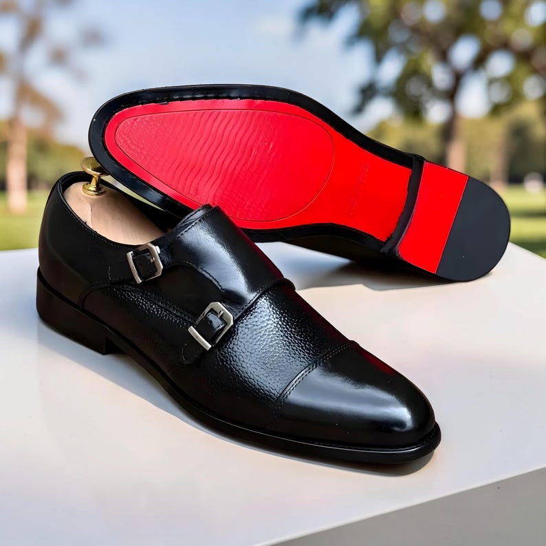Peut inclure: Chaussures de ville noires en cuir avec double bride et boucles argent&eacute;es. La semelle est rouge. Une chaussure est &agrave; l'endroit, l'autre &agrave; l'envers, montrant la semelle rouge. Les chaussures sont sur une surface blanche.