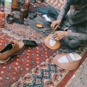 May include: A shoemaker working on a pair of brown leather shoes. The image shows the shoemaker's workspace, including tools, materials, and a partially completed shoe. The shoes have a white accent on the top.