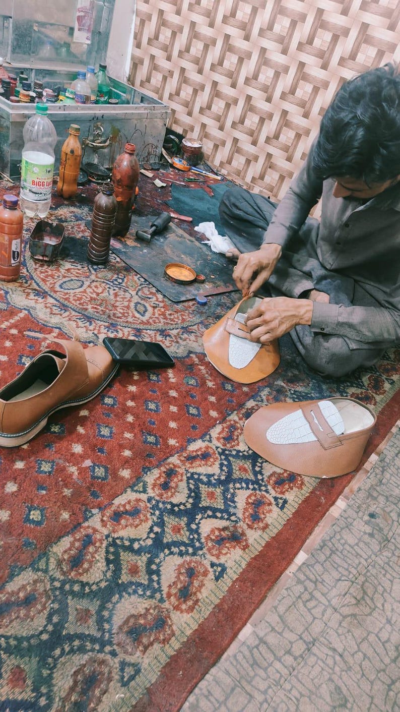 May include: A shoemaker working on a pair of brown leather shoes. The image shows the shoemaker's workspace, including tools, materials, and a partially completed shoe. The shoes have a white accent on the top.
