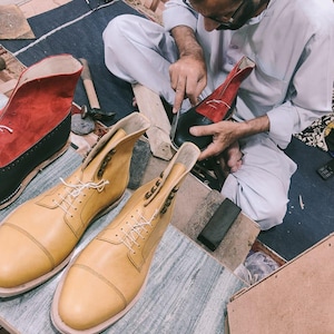 May include: A shoemaker working on a pair of leather boots. The boots are tan and red, with white laces. The shoemaker is using a knife to cut the leather. Other tools and materials are visible.