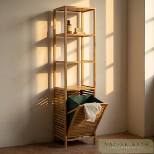 May include: A tall, light-brown bamboo storage unit with open shelves and a slatted laundry hamper. The hamper has a green fabric liner and a white towel. The top shelves hold a bottle and soap. The text "NATIVE BATH CRAFTED PRACTICALLY" is at the bottom.