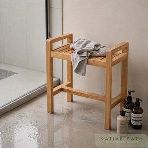 Puede incluir: Un banco de ducha de bambú marrón claro con una parte superior de listones y una toalla gris. El banco tiene forma rectangular y es resistente. La imagen también muestra botellas de productos de baño. El texto "NATIVE BATH CRAFTED PRACTICALLY" está en la parte inferior.
