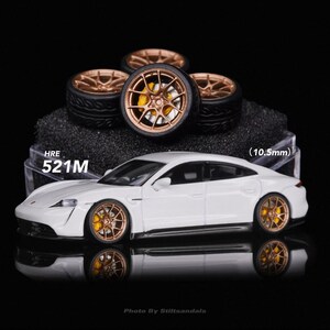 May include: A white model car with bronze-colored wheels and yellow brake calipers. The wheels are displayed separately on a black surface. The text "HRE 521M" is visible. The car's wheels measure 10.5mm.