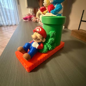 Könnte beinhalten: Eine 3D-gedruckte Figur von Mario, die sich auf einer orangefarbenen Plattform ausruht, neben einem grünen Rohr und einem grünen Busch. Mario trägt seinen roten Hut, blaue Latzhosen und braune Schuhe. Die Figur ist eine farbenfrohe Darstellung des Videospielcharakters.