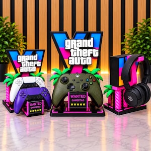 May include: Three black display stands featuring the Grand Theft Auto V logo in blue, pink, and yellow. The stands hold a white and purple game controller, an olive green game controller, and black headphones, each with a Wanted Gamertag sign.