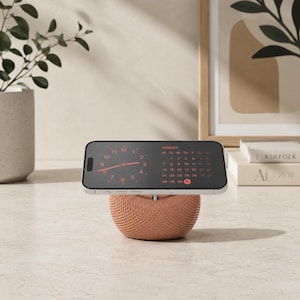 May include: A rose gold, spherical, textured phone stand supporting a smartphone in landscape orientation. The phone screen displays a digital clock and an August calendar with red text on a black background, showing the date 31 highlighted in red.