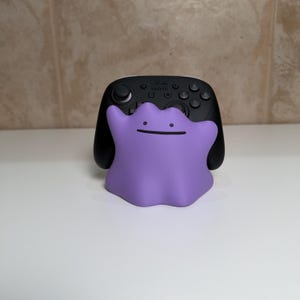 May include: A black Nintendo Switch Pro Controller with a purple Ditto figure in front. The Ditto figure has a simple smiling face. The controller has buttons and joysticks.