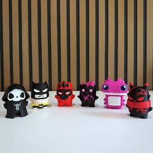 May include: A set of six small, stylized vinyl character figurines arranged in a row. The figures include a black-hooded skeleton, a black and white superhero, a red and black horned creature, a black and pink monster, a pink dragon, and a black and red demon.