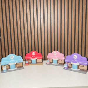May include: Four miniature Pokémon Center building models arranged in a row. Each structure features a tan base, a central blue door, and a roof in light blue, red, pink, or purple. Each building includes a small matching colored kiosk.