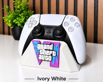 May include: A white gaming controller stand featuring a pink and blue Grand Theft Auto VI logo on the front. The stand is designed to hold a white and black wireless game controller. The text Ivory White is printed below the stand.