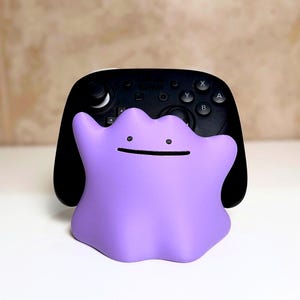 May include: A purple Ditto figure with a simple face is in front of a black video game controller. The controller has white buttons and a black body. The background is a neutral colour.