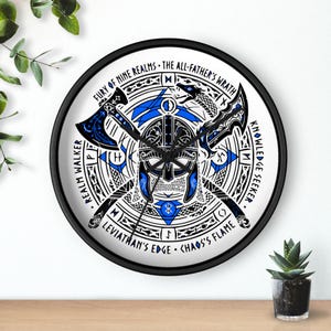 Puede incluir: Un reloj de pared redondo con marco negro y esfera blanca, decorado con ilustraciones gráficas en azul y negro de un casco nórdico, dos hachas cruzadas y una serpiente. El texto dice Fury of Nine Realms, The All-Father's Wrath, Realm Walker, Knowledge Seeker, Leviathan's Edge y Chaos's Flame.