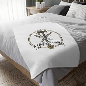 Könnte beinhalten: Eine weiße Fleecedecke mit einem zentralen kreisförmigen Emblem, das eine Doppelaxt und zwei gekreuzte Schwerter zeigt. Unter den Waffen befindet sich ein Banner mit dem Text SERO SED SERIO und einem kleinen Schild mit Löwensymbol.