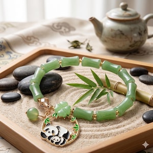 May include: A green jade bracelet with bamboo-shaped beads and gold accents. The bracelet features a charm with a panda bear design. The bracelet is displayed on a sand-filled tray with decorative stones and a teapot.