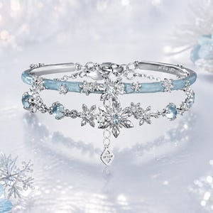 Puede incluir: Dos pulseras plateadas con detalles de copos de nieve y cristales. La pulsera superior tiene una banda azul claro con adornos de cristal. La pulsera inferior presenta un colgante de copo de nieve más grande y pequeños detalles de cristal. Fondo borroso de invierno.
