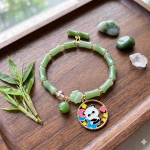 Könnte beinhalten: Ein grünes Jade-Armband mit bambusförmigen Perlen und goldenen Akzenten. Das Armband hat einen Charm mit einem Panda-Bären-Design. Das Armband wird auf einem Holztablett mit Bambusblättern und dekorativen Steinen präsentiert.