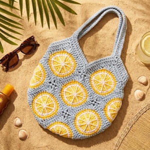 May include: A light blue crochet tote bag with yellow and white lemon slice designs. The bag has two handles and is lying on sand next to sunglasses, sunscreen, and a glass of lemonade. The bag is perfect for a day at the beach.