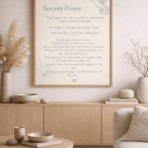Op de afbeelding: Een ingelijste print van het Serenity Prayer, met een duifillustratie en elegant schrift. Het kunstwerk wordt tentoongesteld boven een houten kast, aangevuld met decoratieve vazen, kommen en een salontafel, waardoor een sereen en minimalistisch interieurontwerp ontstaat.