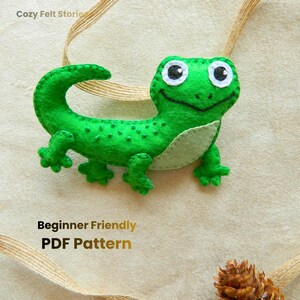 May include: A handmade green felt lizard with white eyes and a smiling mouth. The lizard has a light green belly and is attached to a tan ribbon. Text on the image reads "Beginner Friendly PDF Pattern".