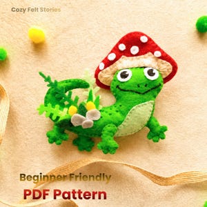 May include: A felt craft project featuring a bright green lizard wearing a red mushroom cap with white spots. The lizard has a light green belly, black eyes, and is decorated with small green felt leaves, yellow pom-poms, and grey felt stones.