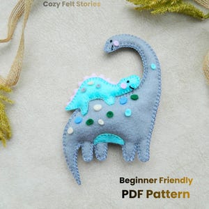 May include: A gray felt dinosaur with a smaller turquoise dinosaur on its back. The dinosaurs have decorative stitching and colorful accents. The text "Cozy Felt Stories" and "Beginner Friendly PDF Pattern" are also visible.
