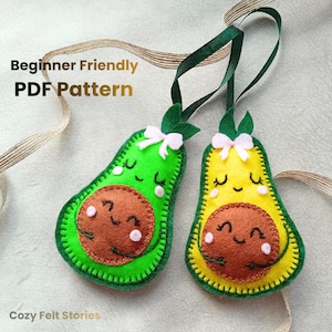Può includere: Due ornamenti a forma di avocado in feltro, uno verde e uno giallo, ciascuno con un seme marrone e una faccia sorridente. Hanno nastri verdi per appenderli e piccoli fiocchi rosa. Il testo "Beginner Friendly PDF Pattern" è visibile.