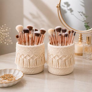 May include: Two cylindrical off-white woven rope baskets with fringe detailing, each filled with a collection of makeup brushes featuring rose gold ferrules and light-colored bristles, arranged on a smooth surface with gold jewelry nearby.