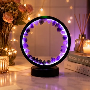 May include: A black circular LED lamp featuring a ring of purple amethyst crystals. The lamp has a black base and emits a soft purple light. This decorative lamp is a home accent.