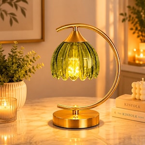 Pode incluir: Uma luminária de mesa decorativa com cúpula de vidro verde e base e braço curvo de metal dourado. A luminária tem uma cúpula com bordas e um brilho quente. A luminária está sobre uma superfície semelhante a mármore.