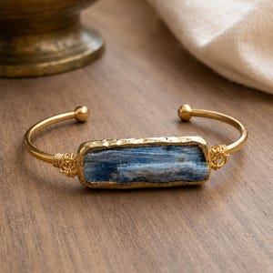 May include: A gold-toned cuff bracelet featuring a rectangular, raw blue kyanite stone set in a textured gold-colored bezel. The metal band is accented with intricate wire-wrapped detailing on both sides of the central stone and ends in small spheres.