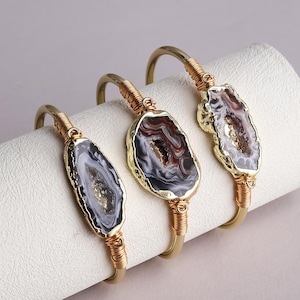 Puede incluir: Tres pulseras de alambre en tono dorado, cada una con una rodaja de ágata cruda central. Las piedras de ágata muestran bandas concéntricas de gris, blanco y marrón con centros cristalinos. El alambre metálico envuelve la banda cerca de la piedra.