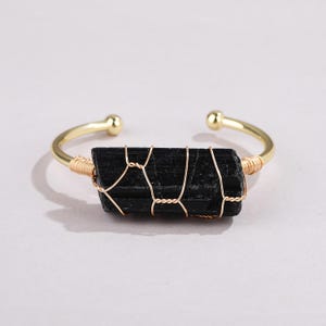 May include: A gold-toned metal cuff bracelet featuring a raw, rectangular black tourmaline crystal at the center. The crystal is secured to the open-ended band with thin, decorative gold-colored wire wrapping that crisscrosses over the stone.