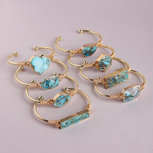 May include: A collection of seven gold-tone wire cuff bracelets, each featuring a unique stone inlay. The stones display various shades of blue and turquoise with brown veining. The shapes include hearts, rectangles, ovals, and hexagons.