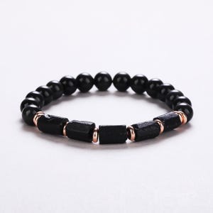 Könnte beinhalten: Ein elastisches Armband mit einer Reihe glatter, runder schwarzer Perlen und fünf länglichen, rau strukturierten schwarzen Zylinderperlen. Die Zylinderperlen sind durch kleine, polierte roségoldfarbene Metallzwischenringe getrennt.