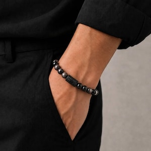 May include: A beaded bracelet featuring smooth, dark, reflective spherical beads and a single rectangular, textured black centre bead. The bracelet is worn on a wrist, contrasting against a black long-sleeved shirt and black trousers.
