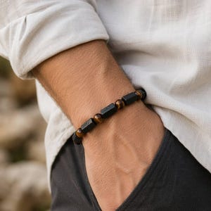 Könnte beinhalten: Ein Armband mit abwechselnden schwarzen rechteckigen Perlen und runden braunen Tigerauge-Perlen. Das Armband wird am Handgelenk getragen und zeigt ein sich wiederholendes Muster aus dunklen, matt texturierten rechteckigen Segmenten, die durch glatte, polierte, goldbraune kugelförmige Steine getrennt sind.