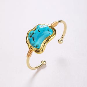 May include: A gold-tone cuff bracelet featuring a central, irregular, bright blue turquoise stone with dark matrix veining. The stone is encased in a textured gold-coloured metal frame, with thin gold wire wrapping around the band on both sides.