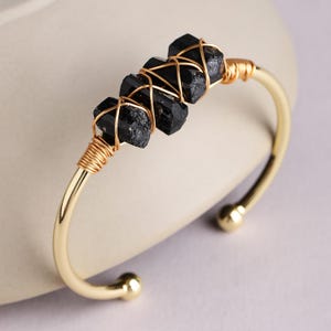Puede incluir: Una pulsera tipo brazalete de tono dorado con tres cristales negros en bruto de forma columnar, sujetos al centro de la banda con un fino alambre dorado enrollado. La pulsera tiene un acabado liso y pulido con extremos esféricos.