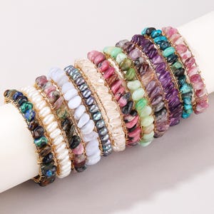 Op de afbeelding: Een collectie van elf met draad omwikkelde armbanden met diverse gepolijste edelsteen- en parelkralen. De armbanden tonen een spectrum aan kleuren, waaronder wit, blauw, paars, roze, groen en donkergrijs, elk vastgezet met dun goudkleurig draad.