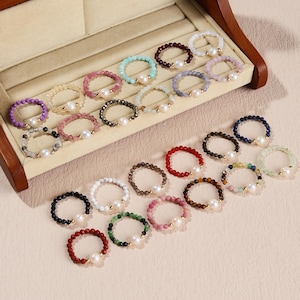 Puede incluir: Una colección de anillos de cuentas en varios colores, incluyendo morado, rosa, verde y marrón. Cada anillo presenta un acento de perla central. Los anillos se exhiben en una caja de joyería de madera.