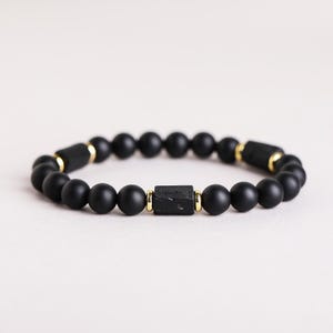 May include: A beaded bracelet featuring matte black spherical beads interspersed with three rectangular black textured beads. Each rectangular bead is flanked by small, polished gold-tone metal spacers, creating a repeating pattern along the circular band.