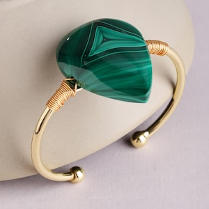 May include: A gold-tone cuff bracelet featuring a large, teardrop-shaped malachite stone with distinct dark and light green banding. The stone is secured to the polished metal band by coiled gold-colored wire wraps on both sides of the setting.