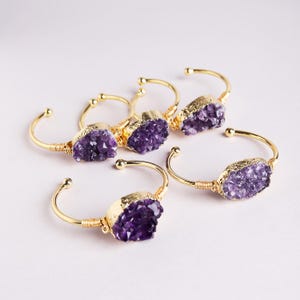 May include: A collection of five open cuff bracelets featuring raw, deep purple amethyst crystal clusters. Each bracelet is crafted from gold-toned wire that wraps around the base of the stone, creating a polished and structured look for each piece.