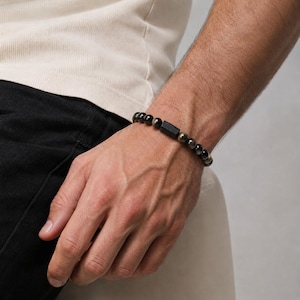 May include: A beaded bracelet featuring smooth, round black and metallic gold-toned beads arranged in a circular pattern, centred with a single rectangular black bead. The bracelet is constructed with a flexible cord to fit around the wrist.